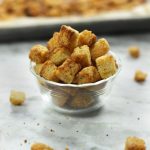 A small clear glass bowl full of Sourdough Croutons sitting on a white marble countertop.