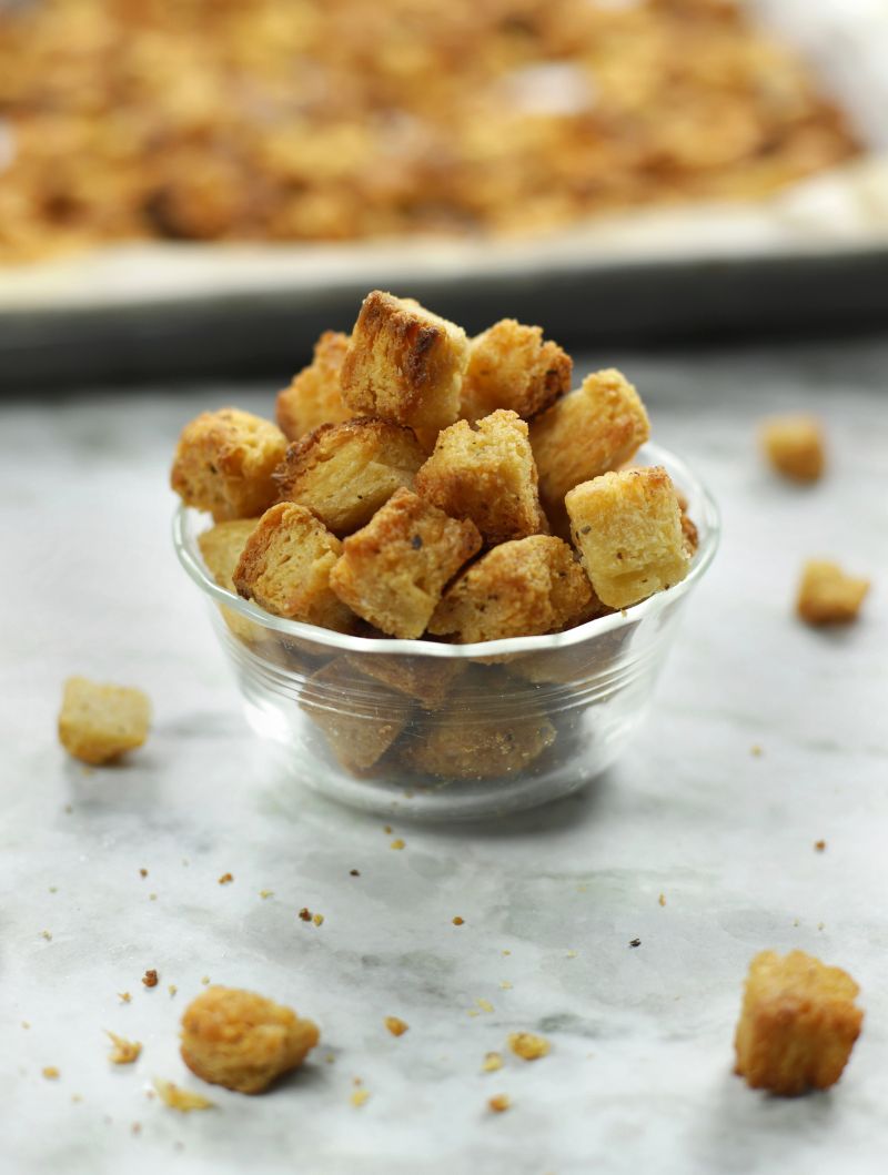 A small clear glass bowl full of Sourdough Croutons sitting on a white marble countertop.