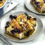 Golden Sourdough Bread Casserole with blueberries and maple syrup sits on gray plates atop a white marble surface.